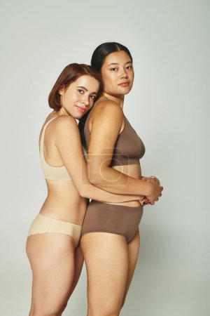 two multiethnic women in underwear embracing and looking at camera on light grey background
