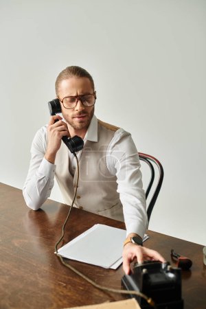 Photo for Good looking young man with glasses and beard sitting at table and talking by retro phone in office - Royalty Free Image