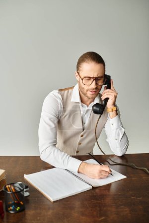 handsome bearded man with glasses talking by retro phone and taking notes at table in office