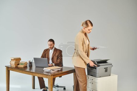 Photo for Young attractive woman in elegant suit using copy machine while her boyfriend working on laptop - Royalty Free Image