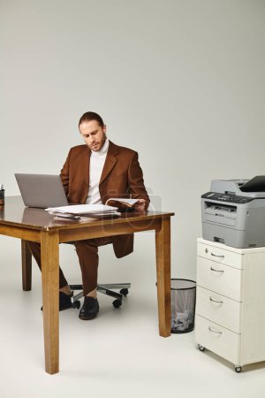 attraktiver bärtiger Mann in brauner schicker Jacke, der an seinem Laptop arbeitet und Papierkram betrachtet