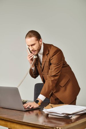 gut aussehender bärtiger Mann im schicken Anzug spricht per Retro-Telefon und schaut im Büro auf seinen Laptop