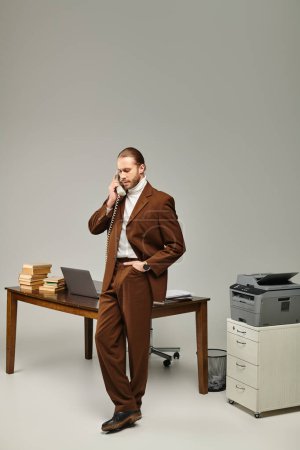 handsome bearded man in brown fashionable jacket talking by retro phone with hand in pocket