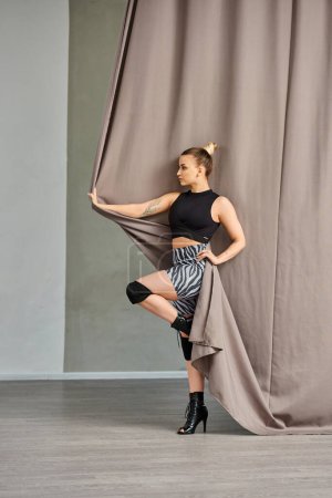 woman strikes a pose in a graceful dance move, balancing on one foot against a curtain-covered wall