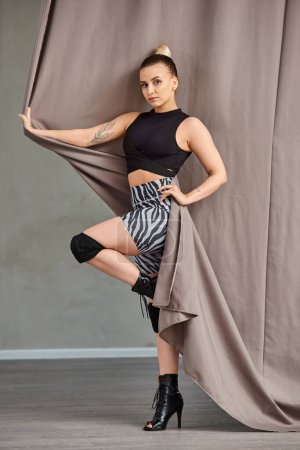 young woman in stylish clothing and high heels gracefully poses against a wall with curtain