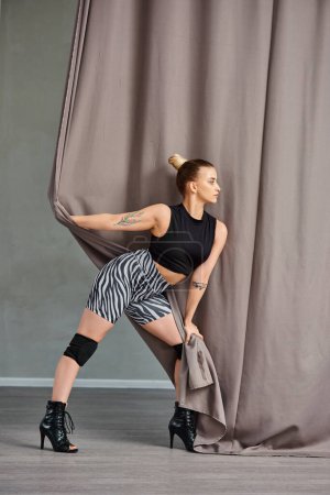 pretty woman in dance clothing and high heels gracefully poses against a wall with curtain