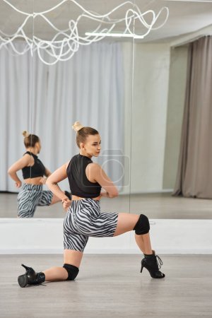 Una bailarina elegante en tacones altos, pantalones cortos de zebra y un top negro, se extiende delante del espejo