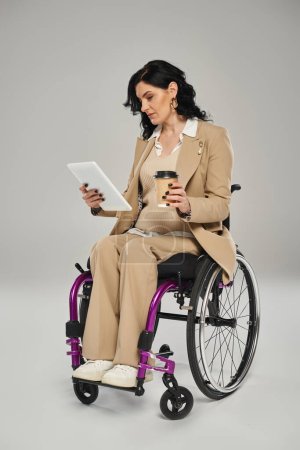 gut aussehende behinderte Frau in pastellfarbener schicker Kleidung im Rollstuhl schaut auf Tablet und hält Kaffee