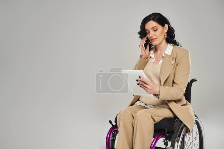 mujer segura de sí misma con discapacidad al mirar la tableta y hablar por teléfono mientras está en silla de ruedas