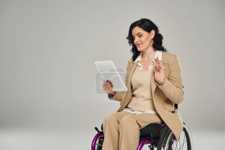 cheerful beautiful disabled woman in pastel attire in wheelchair waving at tablet during video call
