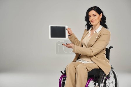 attraktive behinderte Frau im eleganten Anzug sitzt im Rollstuhl und zeigt ihr Tablet vor der Kamera