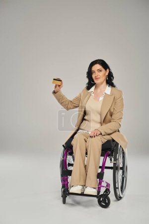 attraktive behinderte Frau im pastellfarbenen Anzug, die im Rollstuhl sitzt und ihre Kreditkarte in die Kamera zeigt