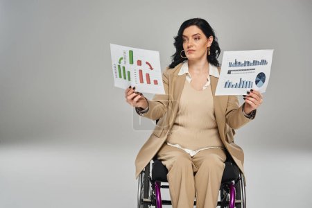 attraktive behinderte Frau mit welligem Haar im schicken Anzug im Rollstuhl beim Betrachten von Grafiken in ihren Händen