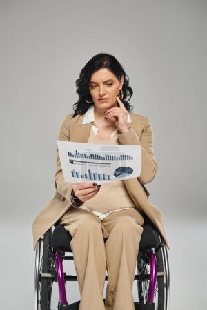 fokussierte behinderte Frau mit welligem Haar im pastellfarbenen Anzug, die im Rollstuhl sitzt und Grafiken betrachtet