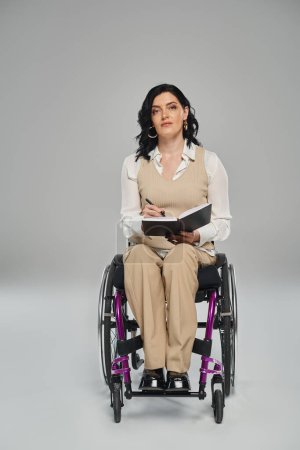 Foto de Atractiva mujer confiada con discapacidad sentada en silla de ruedas con portátil y mirando a la cámara - Imagen libre de derechos