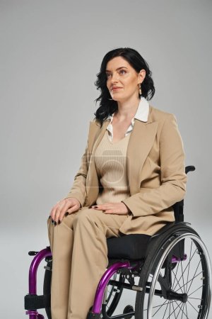 selbstbewusste brünette Frau mit Behinderung im pastellfarbenen Outfit im Rollstuhl sitzend und wegschauend