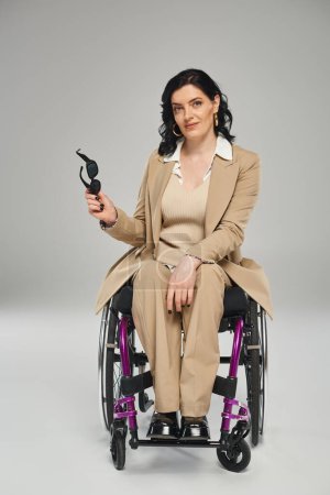 Foto de Hermosa mujer discapacitada en traje pastel en silla de ruedas sosteniendo gafas de sol y mirando a la cámara - Imagen libre de derechos