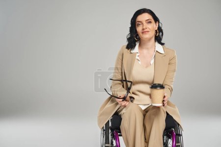 Foto de Atractiva mujer discapacitada en traje pastel sosteniendo gafas de sol y café y mirando a la cámara - Imagen libre de derechos