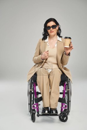 Foto de Mujer confiada con discapacidad en silla de ruedas en gafas de sol sosteniendo café y mirando a la cámara - Imagen libre de derechos