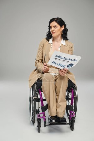 attraktive behinderte Frau im pastellfarbenen Anzug im Rollstuhl, Grafiken und Brille haltend und wegschauend