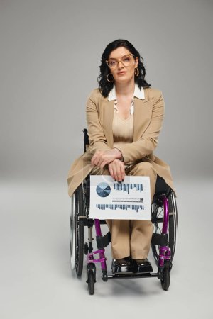 attraktive behinderte Frau mit Brille im Rollstuhl blickt auf Kamera mit Grafik in der Hand
