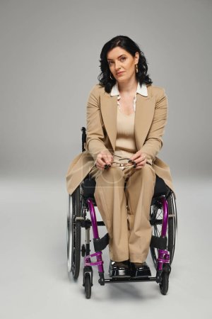 Foto de Mujer segura de buen aspecto en traje de pastel en silla de ruedas sosteniendo gafas y mirando a la cámara - Imagen libre de derechos