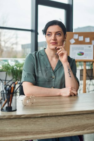 Téléchargez les photos : Séduisante femme d'affaires confiante en tenue décontractée assise à table et regardant loin dans le bureau - en image libre de droit