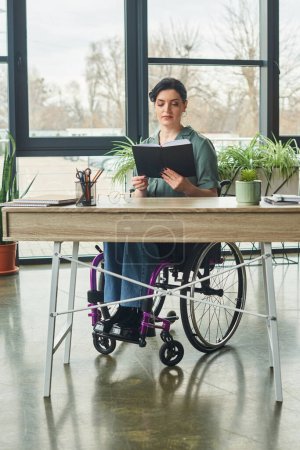 Téléchargez les photos : Bonne regardant femme d'affaires handicapée confiante en fauteuil roulant regardant son carnet dans le bureau - en image libre de droit