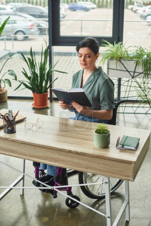 Téléchargez les photos : Femme brune confiante en tenue décontractée en fauteuil roulant travaillant avec son ordinateur portable au bureau - en image libre de droit