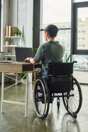 dedicated brunette disabled woman in casual attire sitting in wheelchair and working hard in office