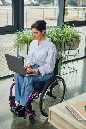 Téléchargez les photos : Femme d'affaires handicapée attentive en tenue décontractée en fauteuil roulant travaillant à l'ordinateur portable pendant son bureau - en image libre de droit