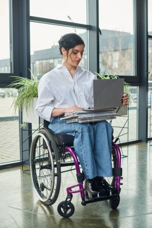 Téléchargez les photos : Femme d'affaires attrayante travaillant dur avec un handicap de mobilité en fauteuil roulant regardant ordinateur portable - en image libre de droit