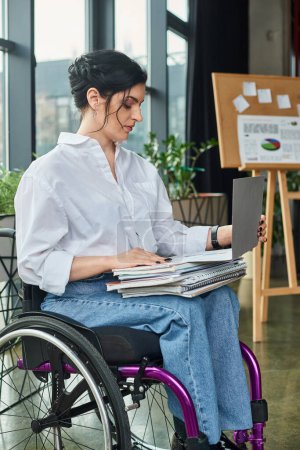 attraktive, hart arbeitende behinderte Geschäftsfrau im Rollstuhl, die mit Papierkram an ihrem Laptop arbeitet