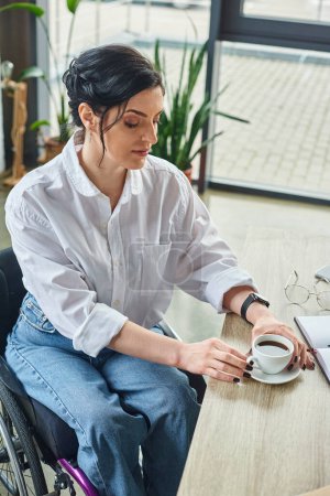attractive disabled woman in stylish attire in wheelchair drinking her coffee while at workplace