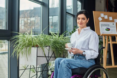 jolly beautiful disabled woman in business attire in wheelchair holding coffee and smiling at camera