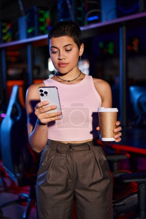 Eine junge Frau steht drinnen, während sie im Cybersport Club ein Telefon und eine Kaffeepapiertasse in der Hand hält.