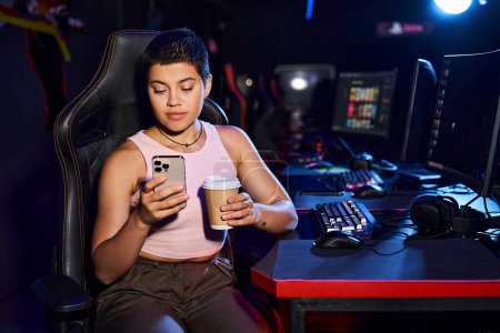 junge stilvolle Frau sitzt an einem Schreibtisch mit einem Telefon in der Hand und einer Tasse Kaffee, Cybersport-Spiele