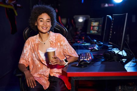 Eine elegante schwarze Frau mit einem Kaffee in der Hand, nimmt sich einen Moment Zeit, um an ihrem Schreibtisch mit Computer zu entspannen