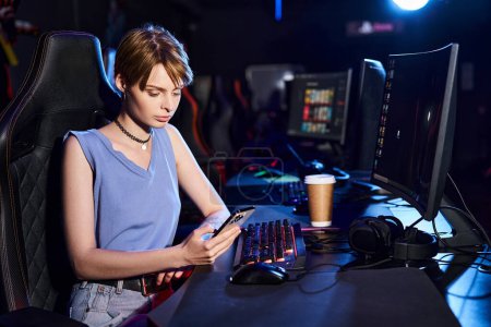 kurzhaarige Frau, die ihr Smartphone in der Nähe des Computers auf dem Schreibtisch benutzt, Cybersport-Spielerin im Computerclub