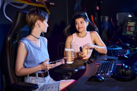 stylish woman holding coffee cup and looking at her female friend, game world and cybersport