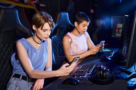Two young women sit at computer desk while scrolling their phones in cybersport game club