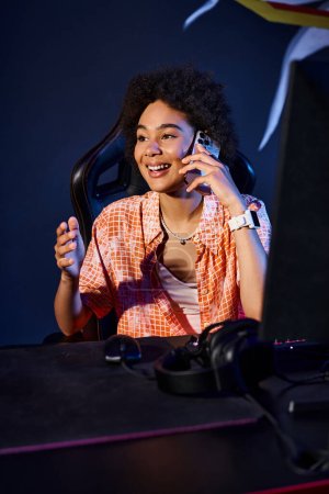 Foto de Alegre negro mujer se sienta en su ordenador y tener llamada telefónica cerca de la computadora, cybersport club - Imagen libre de derechos