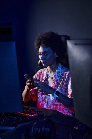 afro-américaine avec les cheveux bouclés en lumière bleue de moniteur d'ordinateur à l'aide de son smartphone