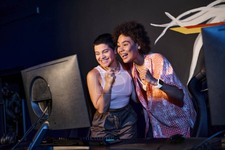 Téléchargez les photos : Deux femmes interraciales et heureuses célébrant la victoire après avoir gagné dans la salle de jeux, cybersport - en image libre de droit