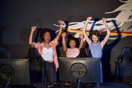 mujeres felices multiculturales mirando el monitor de la computadora y celebrando la victoria, levantaron las manos