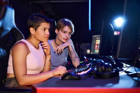 Two women with tattoos concentrating on a cybersport game in computer club room, female gamers