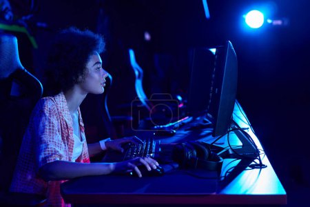Foto de Vista lateral de la mujer afroamericana enfocada que juega intensamente en una sala de luz azul, concepto de ciberdeporte - Imagen libre de derechos