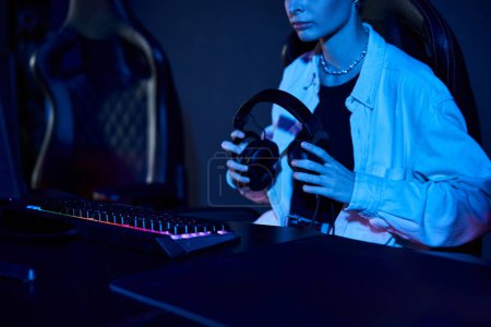 cropped gamer holding headphones and looking at computer in a blue-lit room, cybersport game concept mug #690045488