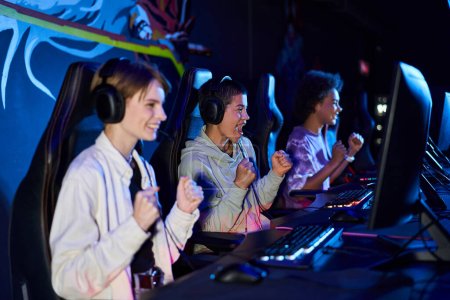 Téléchargez les photos : Excité femmes zoomers interracial engagés dans des jeux cybersport, en utilisant des ordinateurs et des écouteurs - en image libre de droit