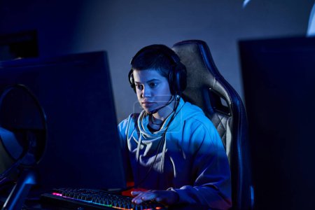 fokussierter Spieler mit kurzen Haaren, der in einem blau beleuchteten Raum auf den Computer schaut, Cybersport-Spieler im Kapuzenpulli